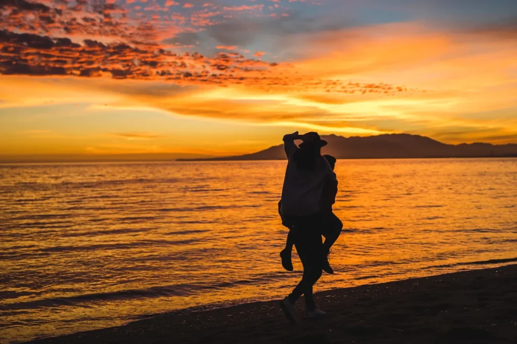 Casal caminhando na praia ao pôr do sol, com céu em tons dourados e mar ao fundo