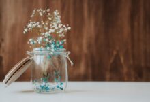 Arranjo delicado de flores azuis e brancas em pote de vidro com tampa de madeira.