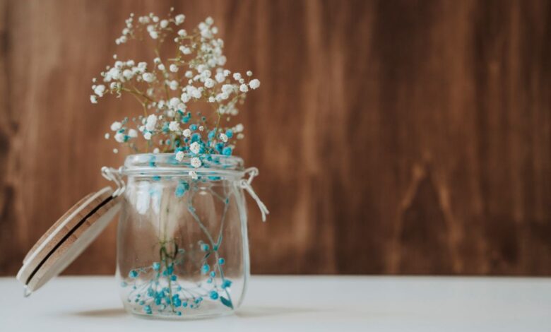 Arranjo delicado de flores azuis e brancas em pote de vidro com tampa de madeira.