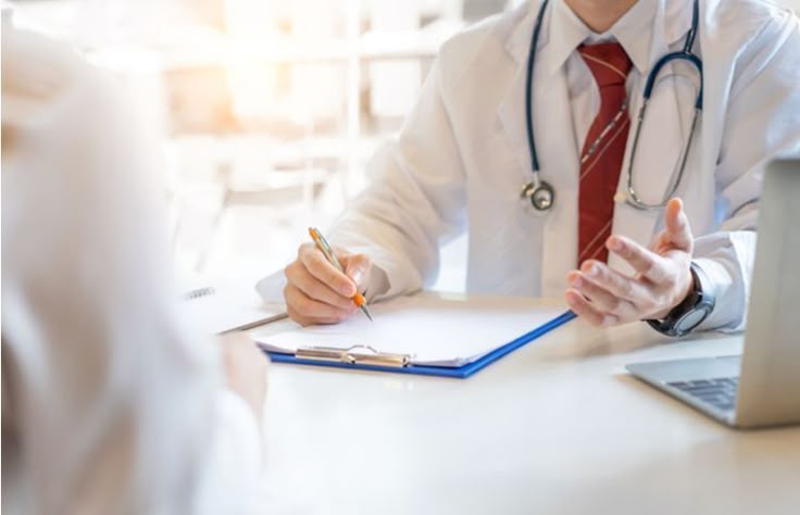 Médico usando jaleco branco conversa com paciente durante consulta, com prancheta e laptop à mesa