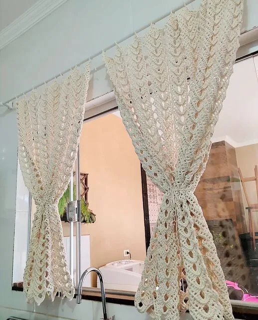 Cortinas de crochê com delicado padrão de ondas, decorando uma janela e trazendo leveza e aconchego ao ambiente.