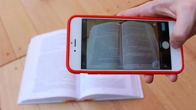 Pessoa fotografando uma página de livro com um smartphone, capturando texto para digitalização.