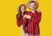Casal sorridente vestindo camisas xadrez vermelhas e chapéus de palha, posando em fundo amarelo.
