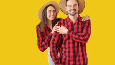 Casal sorridente vestindo camisas xadrez vermelhas e chapéus de palha, posando em fundo amarelo.