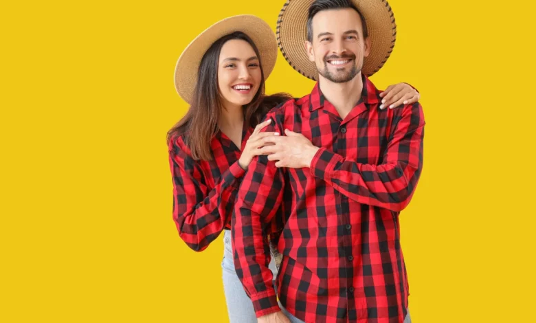 Casal sorridente vestindo camisas xadrez vermelhas e chapéus de palha, posando em fundo amarelo.
