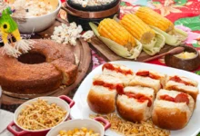 Mesa com comidas típicas de festa junina, incluindo bolo, milho, cachorro-quente, pipoca e amendoim, com decoração festiva.