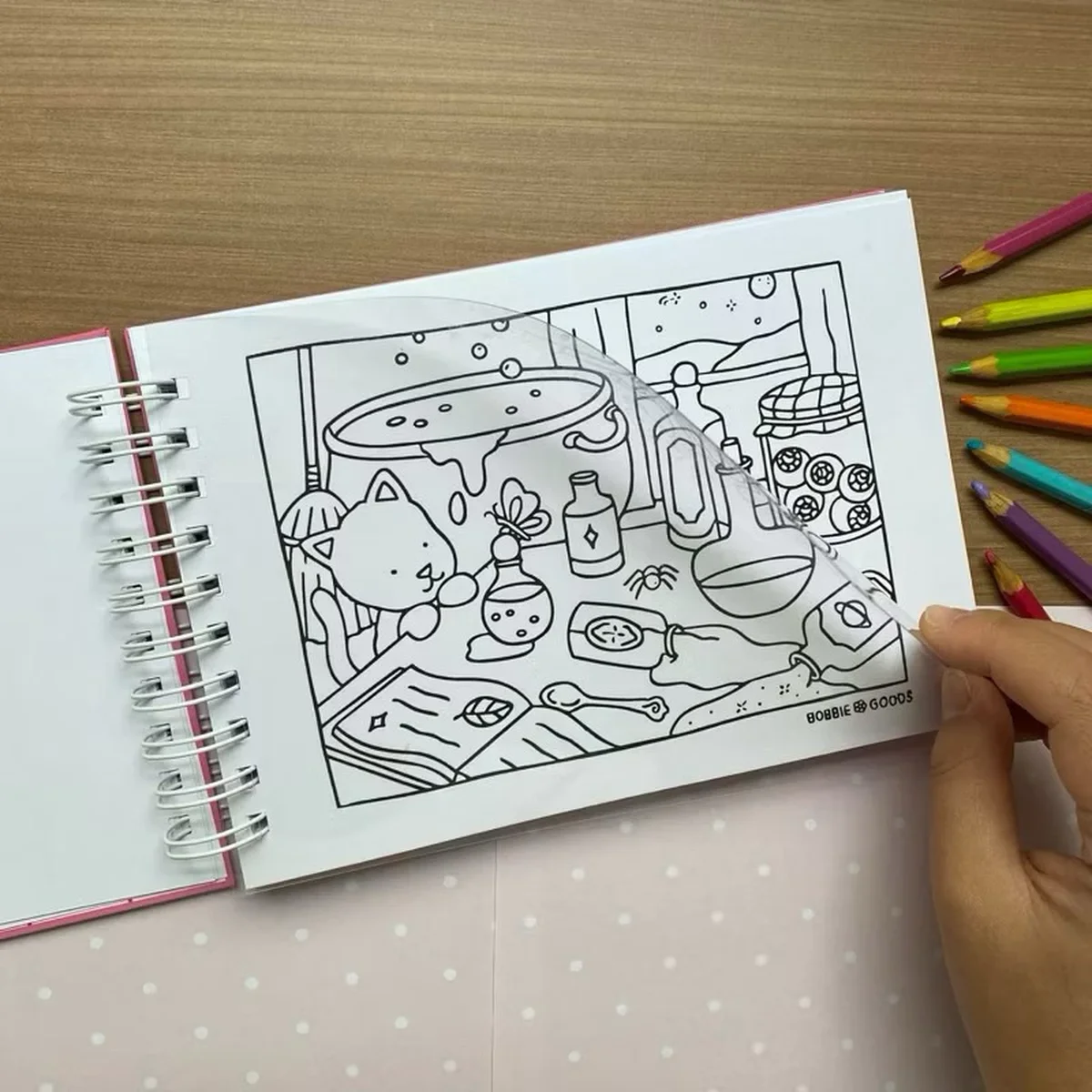 Folha de acetato sendo usada como proteção em livro de colorir Bobbie Goods com lápis ao lado