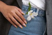 Unhas com francesinha amarela e flor branca no bolso da calça jeans.