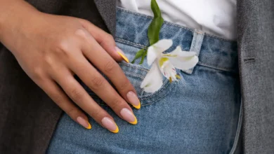 Unhas com francesinha amarela e flor branca no bolso da calça jeans.