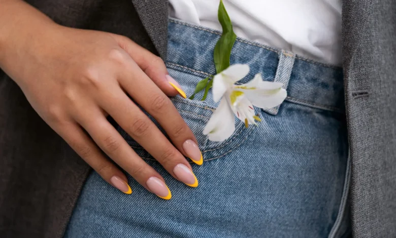 Unhas com francesinha amarela e flor branca no bolso da calça jeans.