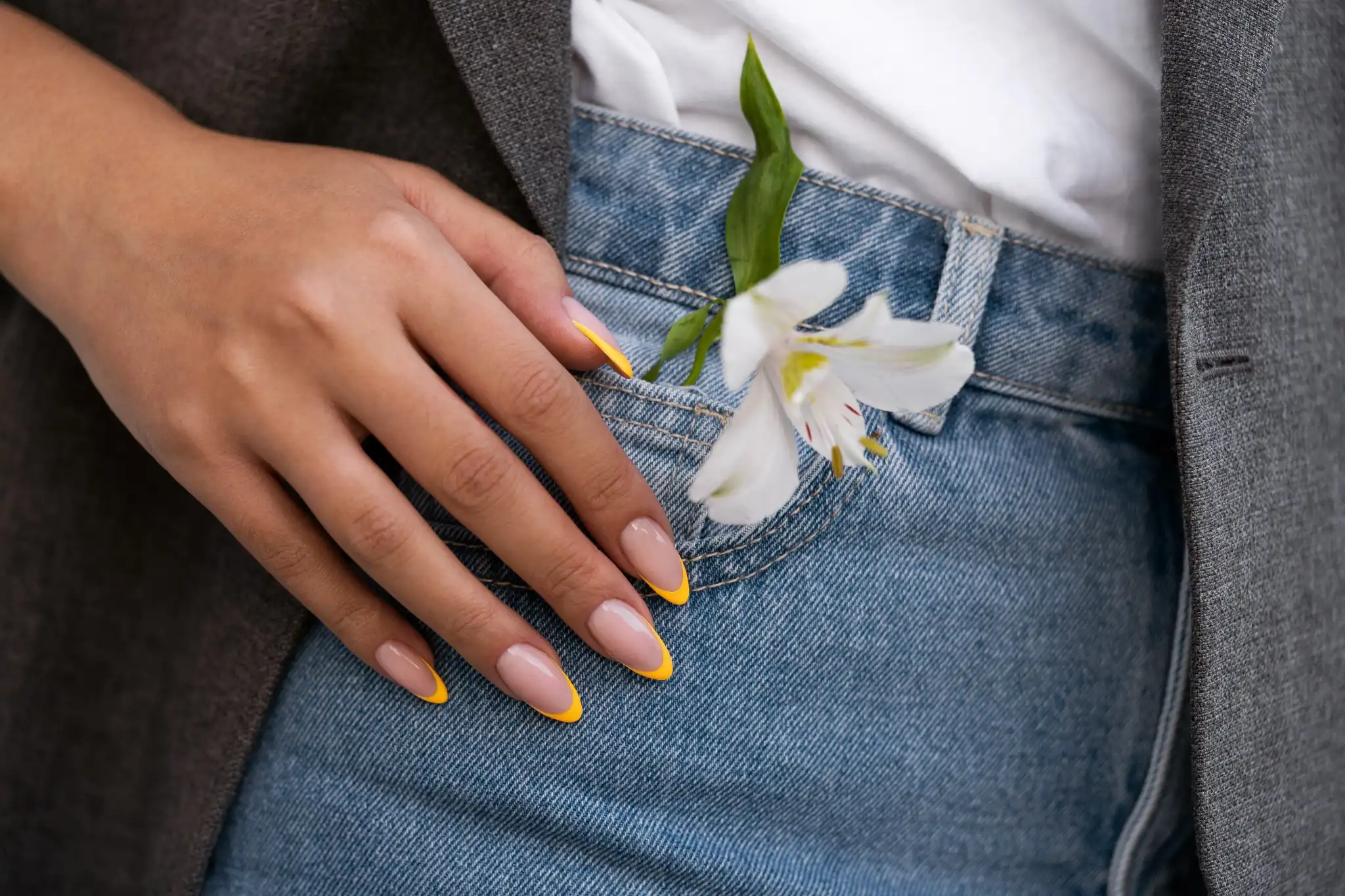 Unhas com francesinha amarela e flor branca no bolso da calça jeans.