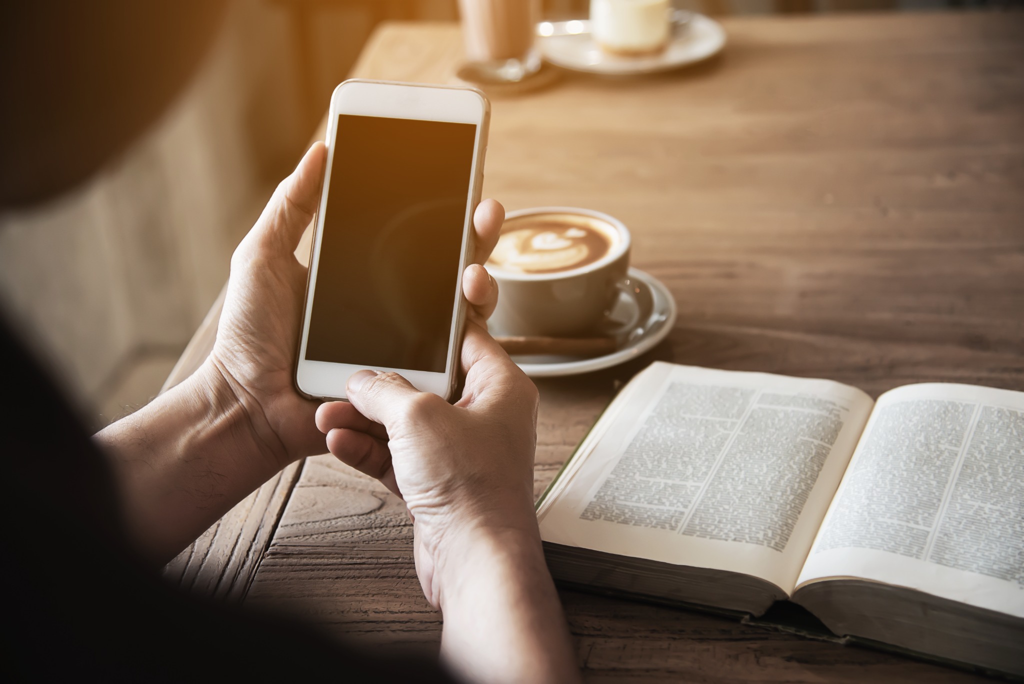 Pessoa usando um smartphone enquanto bebe café e lê uma bíblia em uma mesa de madeira.