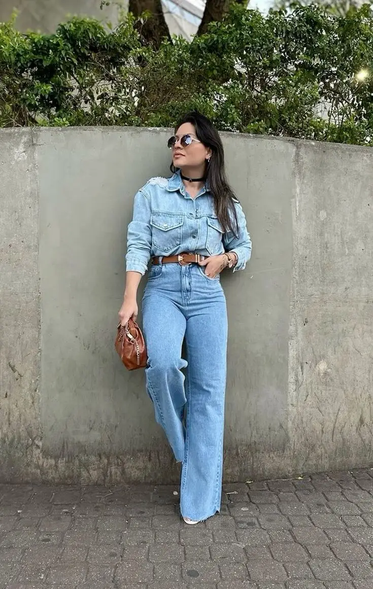 Mulher usando look casual com camisa jeans, calça jeans de cintura alta e bolsa de couro, posando ao ar livre.