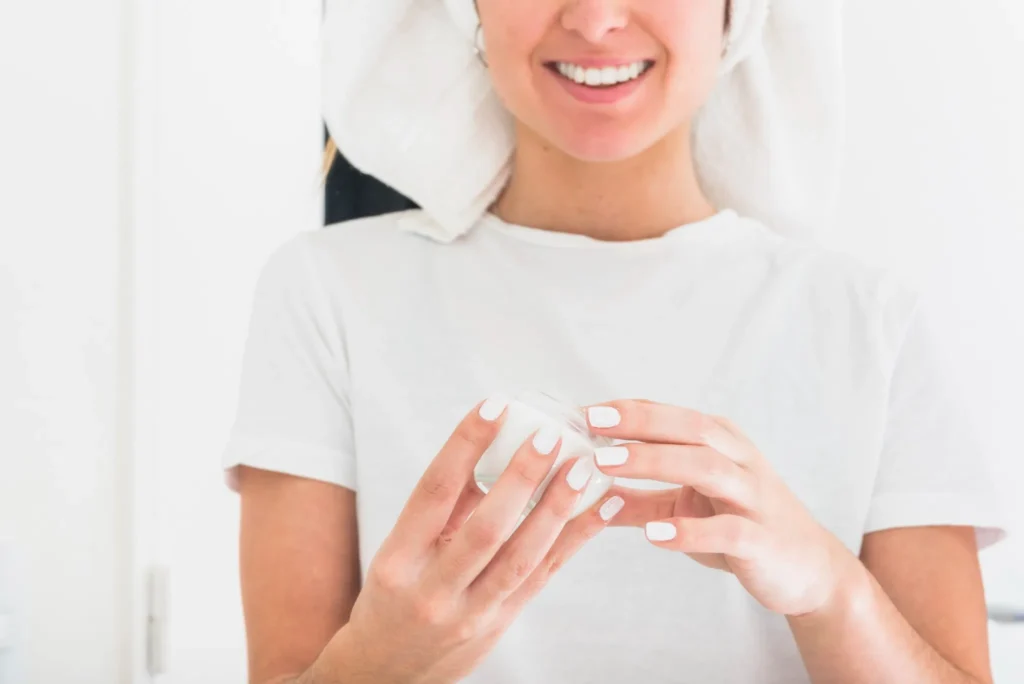 Mulher sorridente com toalha na cabeça segurando pote de creme, pronta para hidratar as mãos antes de dormir.