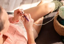 Mulher crochetando com fio branco, criando pontos em um ambiente aconchegante e ensolarado.