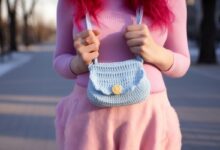 Mulher com cabelo rosa usando uma bolsa de crochê azul com botão amarelo, em ambiente externo.