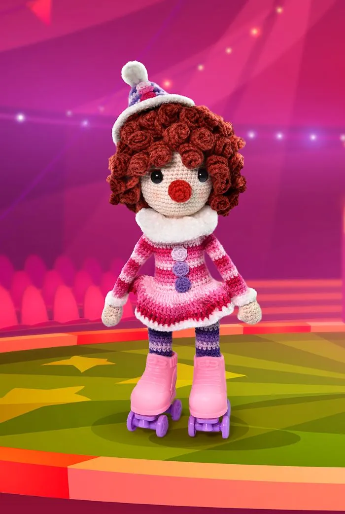Bonequinha de crochê palhacinha com patins rosa, usando um vestido colorido e um chapéu, em um cenário de circo