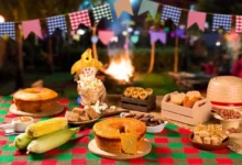 Mesa com comidas típicas de Festa Junina, como bolo de milho, pamonha, paçoca, pipoca e milho verde, com fogueira ao fundo