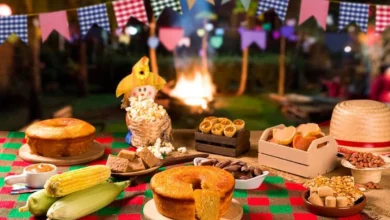 Mesa com comidas típicas de Festa Junina, como bolo de milho, pamonha, paçoca, pipoca e milho verde, com fogueira ao fundo
