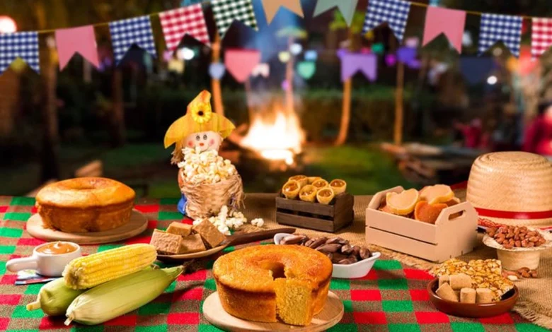 Mesa com comidas tÃpicas de Festa Junina, como bolo de milho, pamonha, paçoca, pipoca e milho verde, com fogueira ao fundo