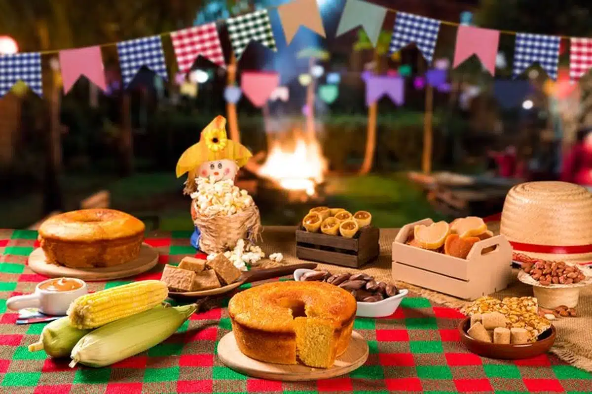 Mesa com comidas típicas de Festa Junina, como bolo de milho, pamonha, paçoca, pipoca e milho verde, com fogueira ao fundo