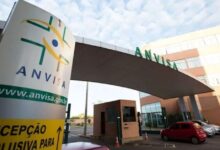 Entrada da sede da Anvisa com placa de recepção e carros estacionados.