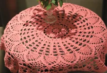 Toalha de crochê rosa sobre mesa redonda com vaso de folhas verdes ao centro