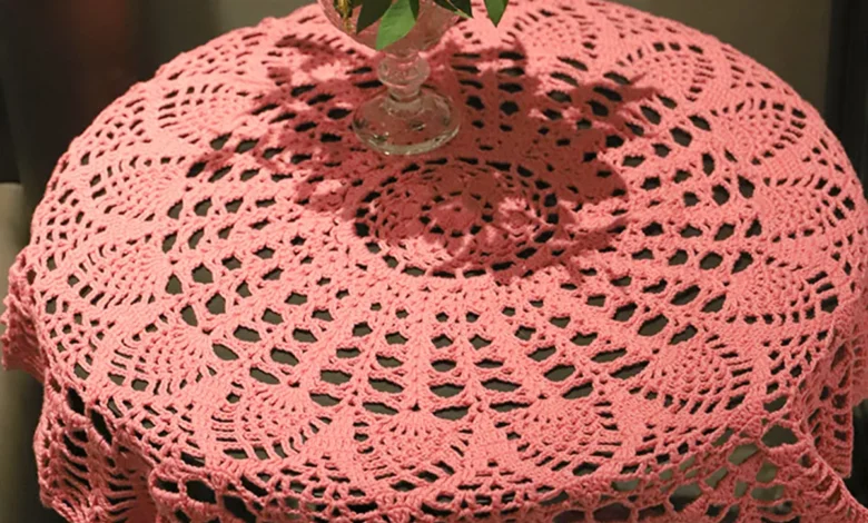 Toalha de crochê rosa sobre mesa redonda com vaso de folhas verdes ao centro