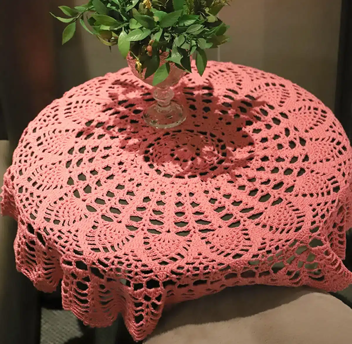 Toalha de crochê rosa sobre mesa redonda com vaso de folhas verdes ao centro