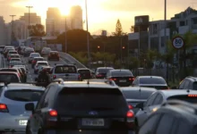 Trânsito intenso ao entardecer em avenida movimentada, com diversos carros parados e sinal vermelho ao fundo.