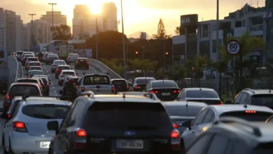 Trânsito intenso ao entardecer em avenida movimentada, com diversos carros parados e sinal vermelho ao fundo.
