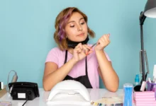 Manicure feminina com camiseta rosa, máscara preta e luvas, cuidando das unhas com alicate, em ambiente de trabalho com equipamentos de manicure.