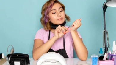 Manicure feminina com camiseta rosa, máscara preta e luvas, cuidando das unhas com alicate, em ambiente de trabalho com equipamentos de manicure.