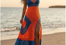 Mulher vestindo vestido longo de crochê laranja e azul com fenda lateral, em cenário de praia ao pôr do sol.