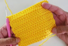 ãos segurando amostra de crochê amarelo vibrante com ponto texturizado, mostrando técnica de crochê fácil com agulha rosa e fio de algodão.