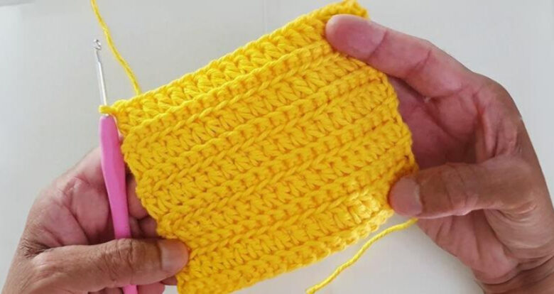 ãos segurando amostra de crochê amarelo vibrante com ponto texturizado, mostrando técnica de crochê fácil com agulha rosa e fio de algodão.