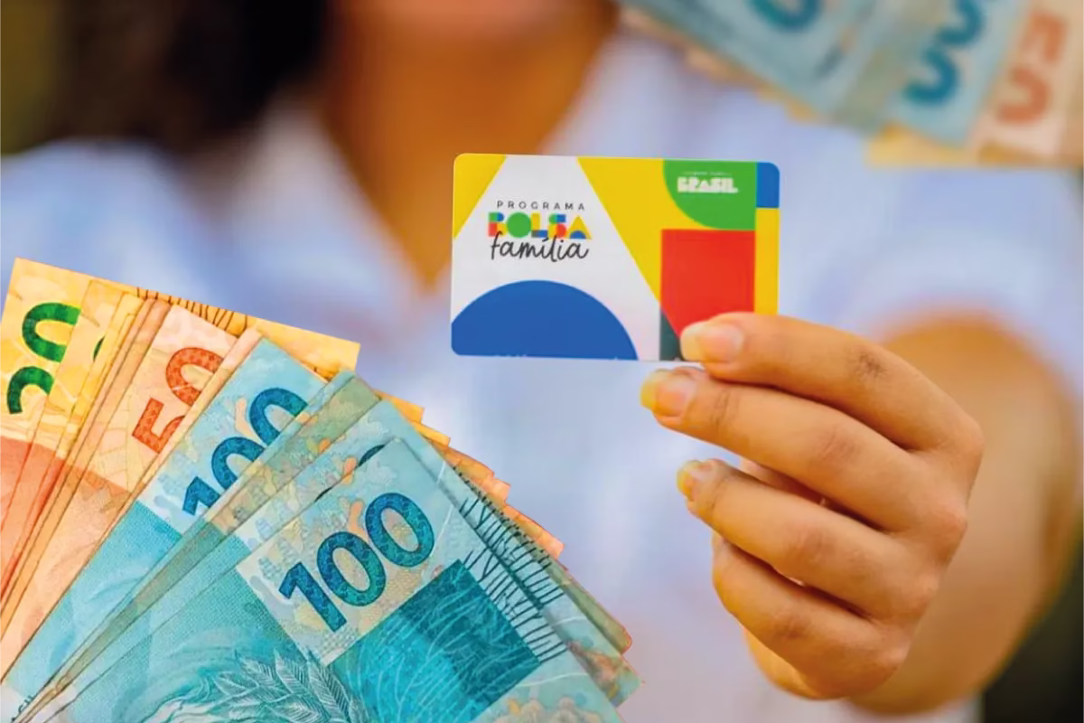 Cartão do programa Bolsa Família sendo segurado, com notas de R$50 e R$100 ao fundo, representando a exclusão de beneficiários após o pente-fino em 2025.