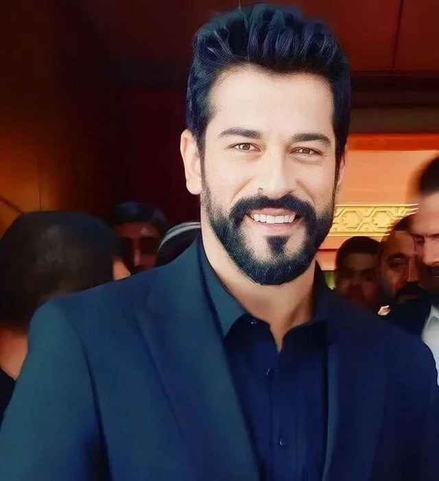 Ator turco Burak Özçivit sorrindo em um evento, com barba e terno preto.