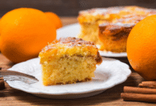 Fatia de bolo de laranja caseiro servida em prato branco com laranjas ao fundo