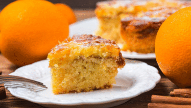 Fatia de bolo de laranja caseiro servida em prato branco com laranjas ao fundo