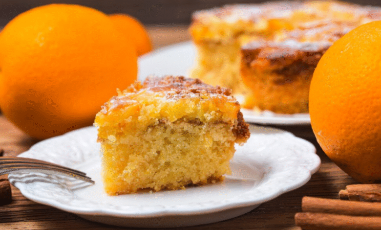 Fatia de bolo de laranja caseiro servida em prato branco com laranjas ao fundo