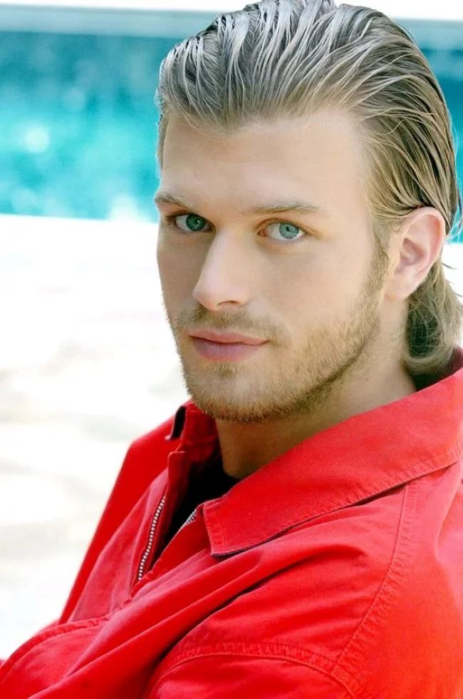 Kıvanç Tatlıtuğ, ator turco de cabelos loiros e olhos azuis, sorrindo em uma foto profissional.
