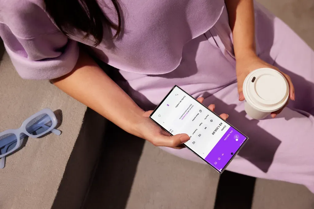 Pessoa usando um smartphone com a interface do aplicativo Nubank enquanto segura um copo, mostrando o design moderno e funcional da conta digital.