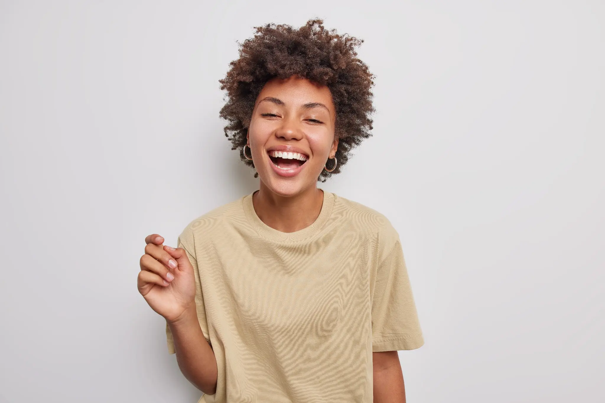 Mulher sorrindo com expressão alegre, usando camiseta bege e cabelo cacheado, sobre fundo branco