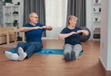 Casal idoso praticando exercícios juntos em casa, sentados em tapete de yoga para manter a saúde e a independência.