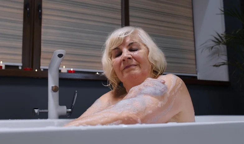 Idosa relaxando em uma banheira, sorrindo enquanto desfruta de um banho reconfortante, com espuma de sabão cobrindo parte do braço.