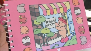Imagem de caderno com desenho fofo de urso segurando uma placa escrita Bobbie Goods em uma loja colorida. Caderno tem personagens de animais ao redor.