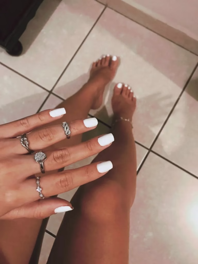 Unhas brancas com acabamento fosco e anéis prateados em primeiro plano, destacando o cuidado profissional.