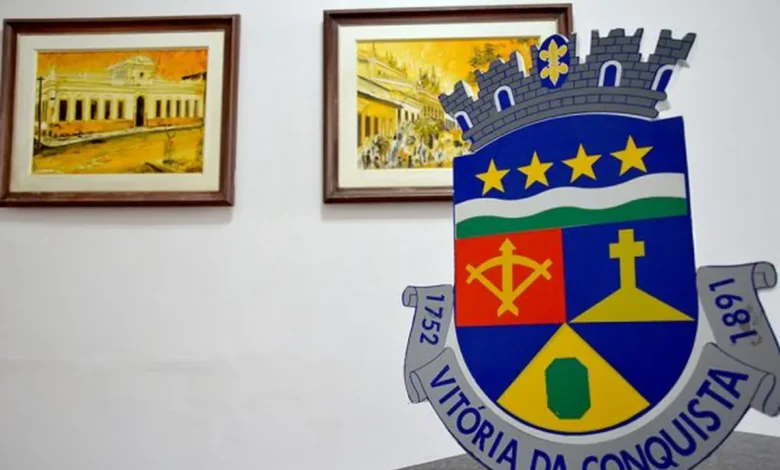 Brasão de Vitória da Conquista, cidade da Bahia, com detalhes de uma cruz, uma espada, e estrelas, no contexto de alerta sobre cadastro habitacional gratuito.