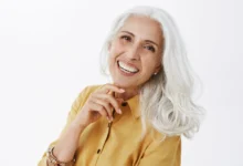 Mulher sorridente com cabelos brancos e elegantes, usando camisa amarela, mostrando confiança e estilo natural.
